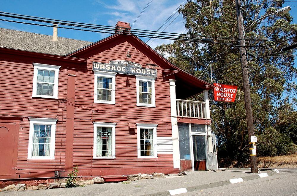 Exterior of old red roadhouse with white trim next to tall pines.