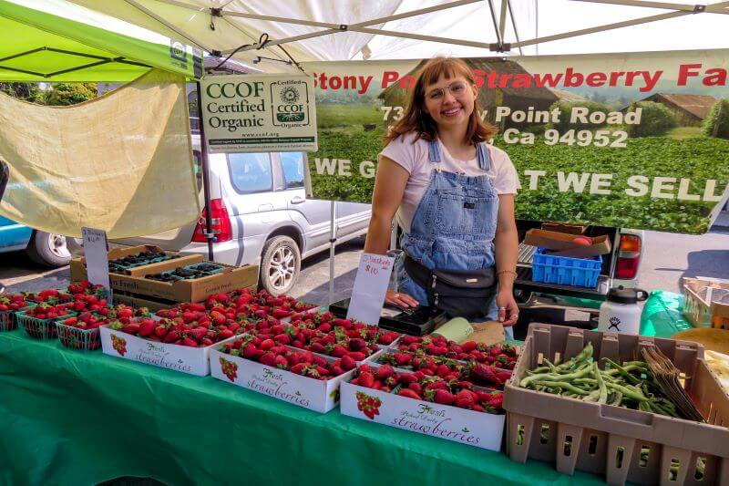Young woman in overalls mans booth at Petaluma Farmers' Market at Walnut Park.