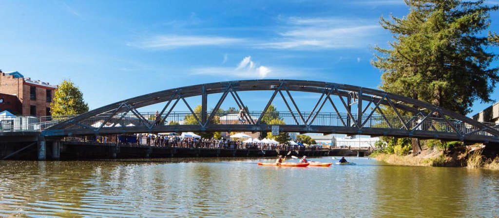 Petaluma River Festivals. People in kayaks on river under bridge, with festival lining waterfront above. 