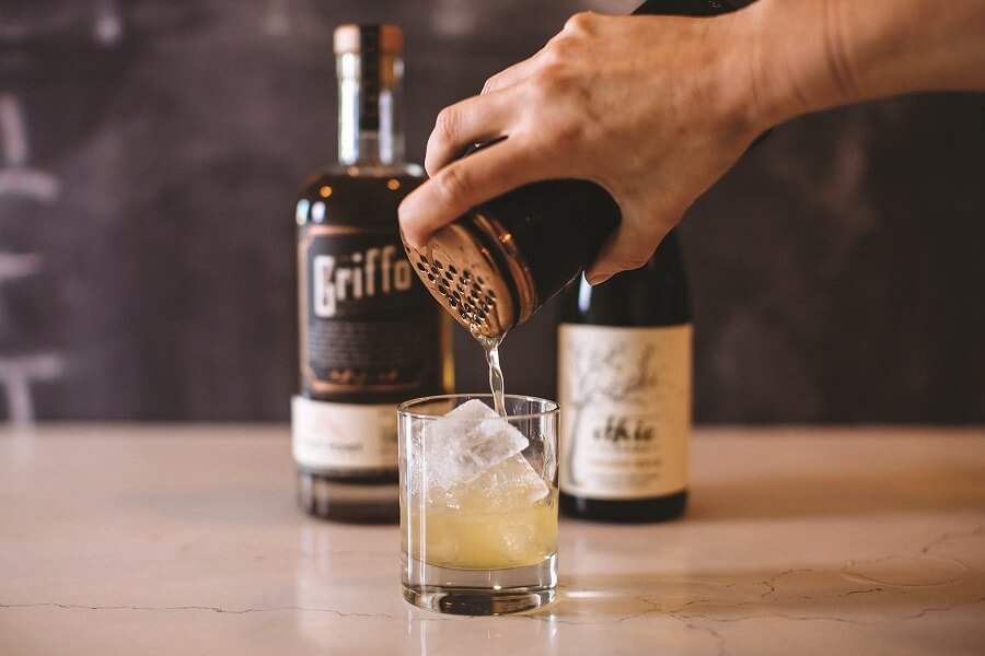 Griffo and other bottle behind hand pouring cocktail from shaker into highball glass