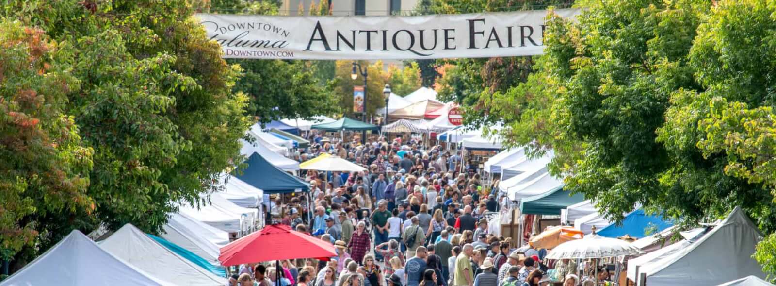 Tree- and tent-lined street filled with hundreds of fair goers. Banner says Downtown Petaluma Antique Fair