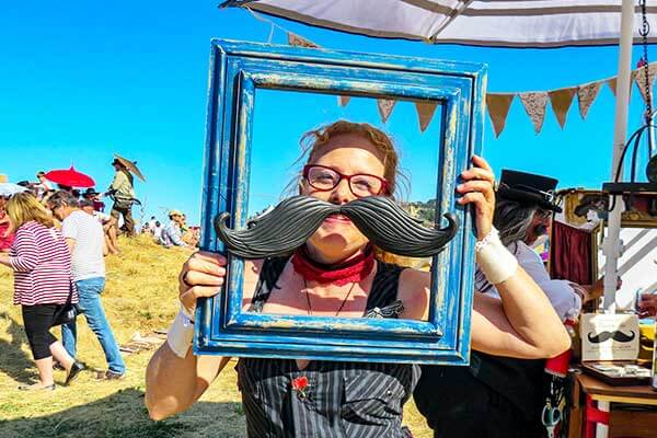 festival_shopping Woman holding a frame with a very large moustache poised over her face at an outdoor festival