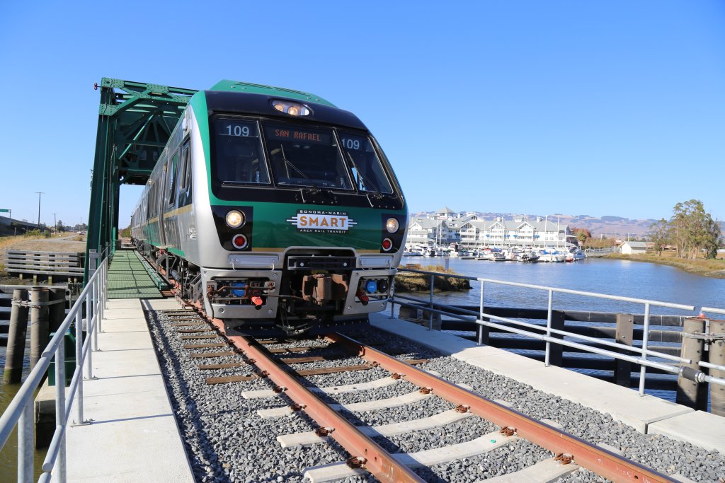 Travel smart to petaluma 5 Train crossing bridge near marina with hotel in background.