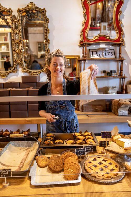 Smiling woman handing bag over a bakery counter with cookies, pie, and muffins.