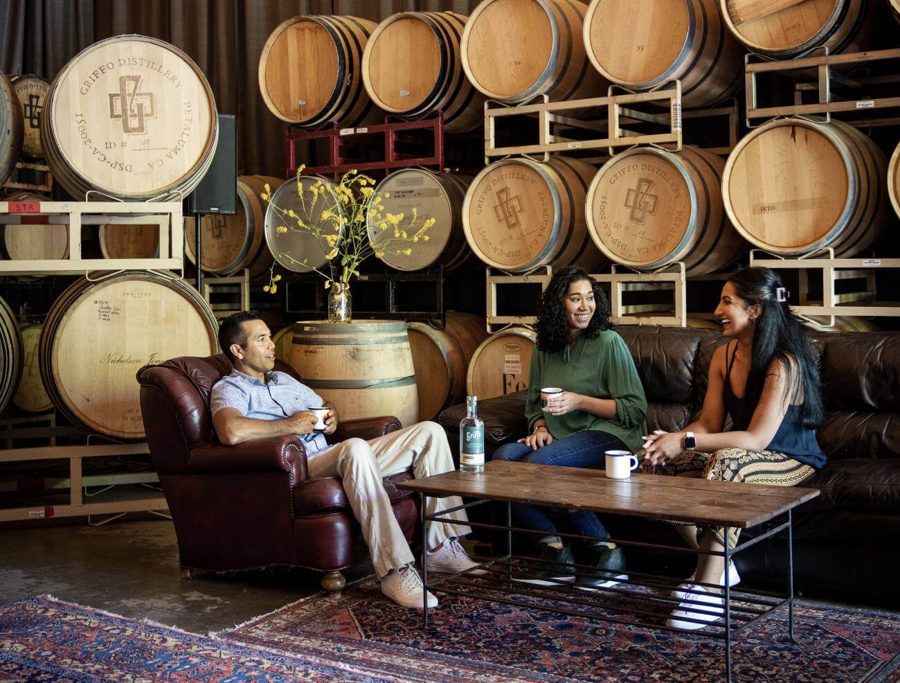Three people on couch and easy chair in barrel room with griffo bottle on coffee table.