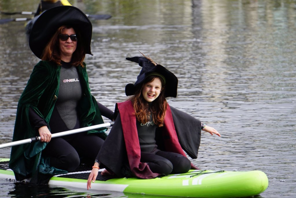 Woman and preteen girl dressed as witches over wetsuits on paddle board.