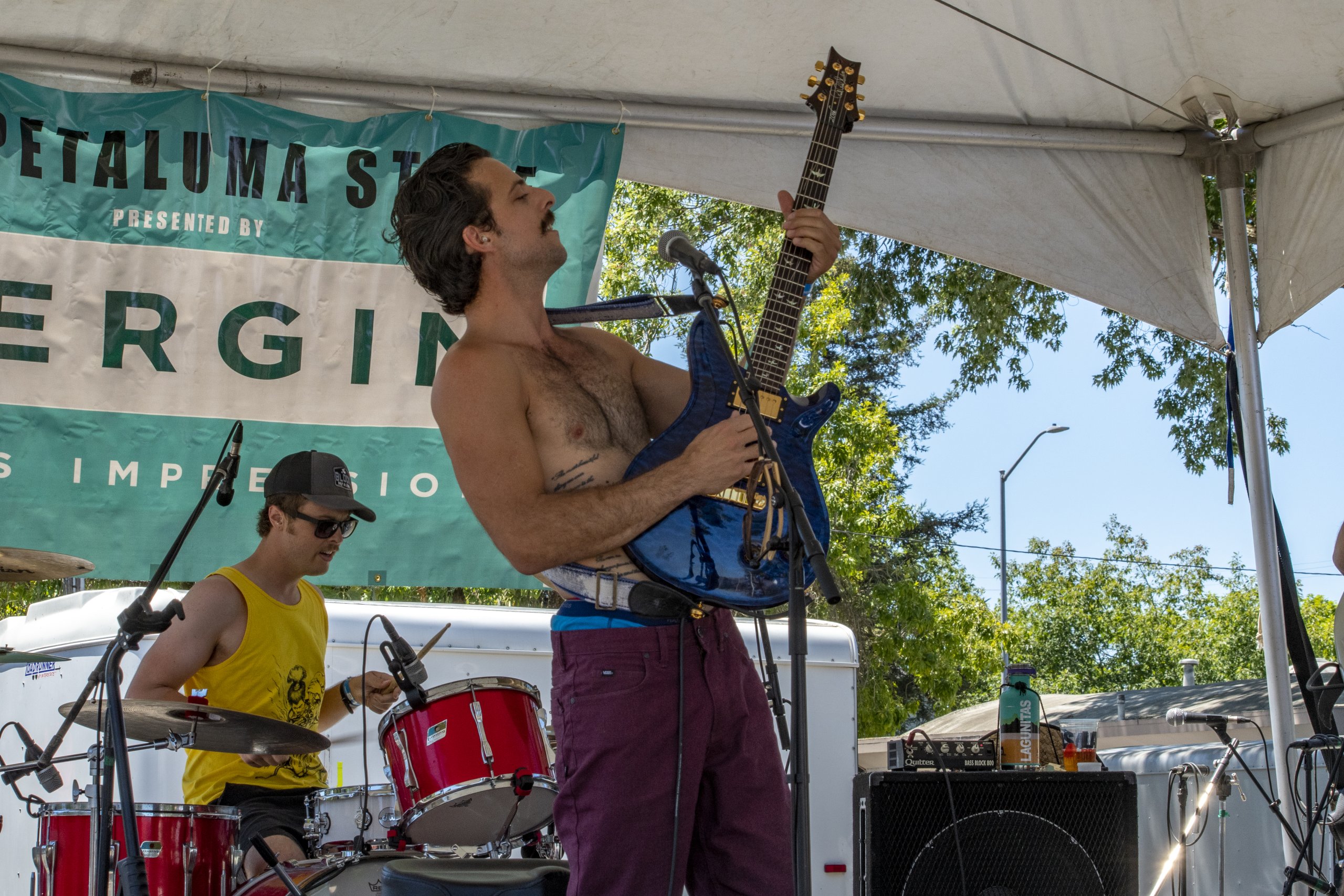 Shirtless lead guitar player performing on outdoor stage, drummer in background.