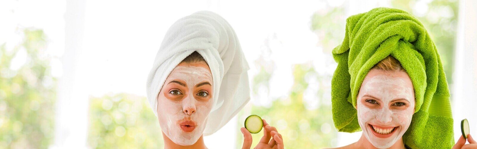 Women with towels on their heads and facial masks on holding cucumber slices up.