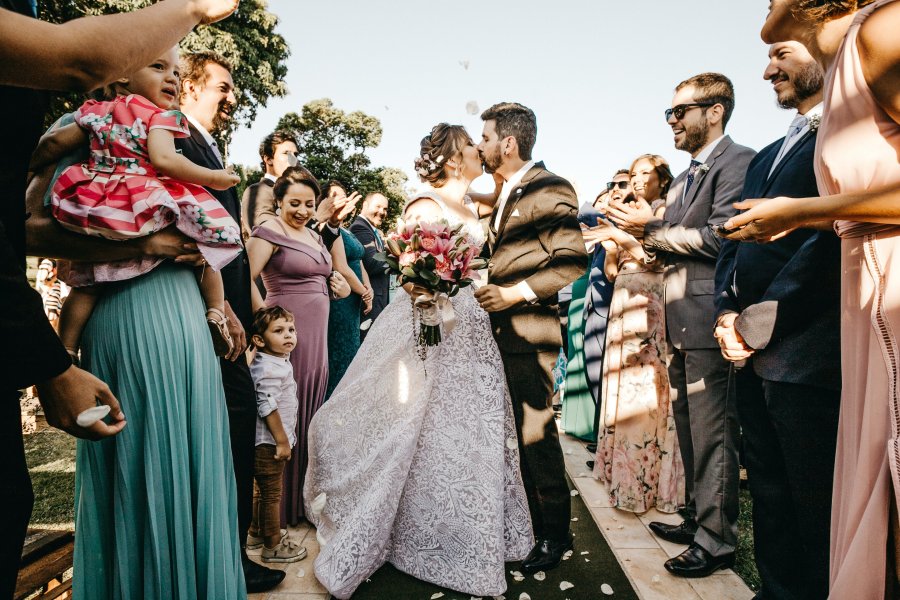 Bride in lace gown holding bouquet to side while kissing groom with guests on either sides clapping, smiling at outdoor wedding.