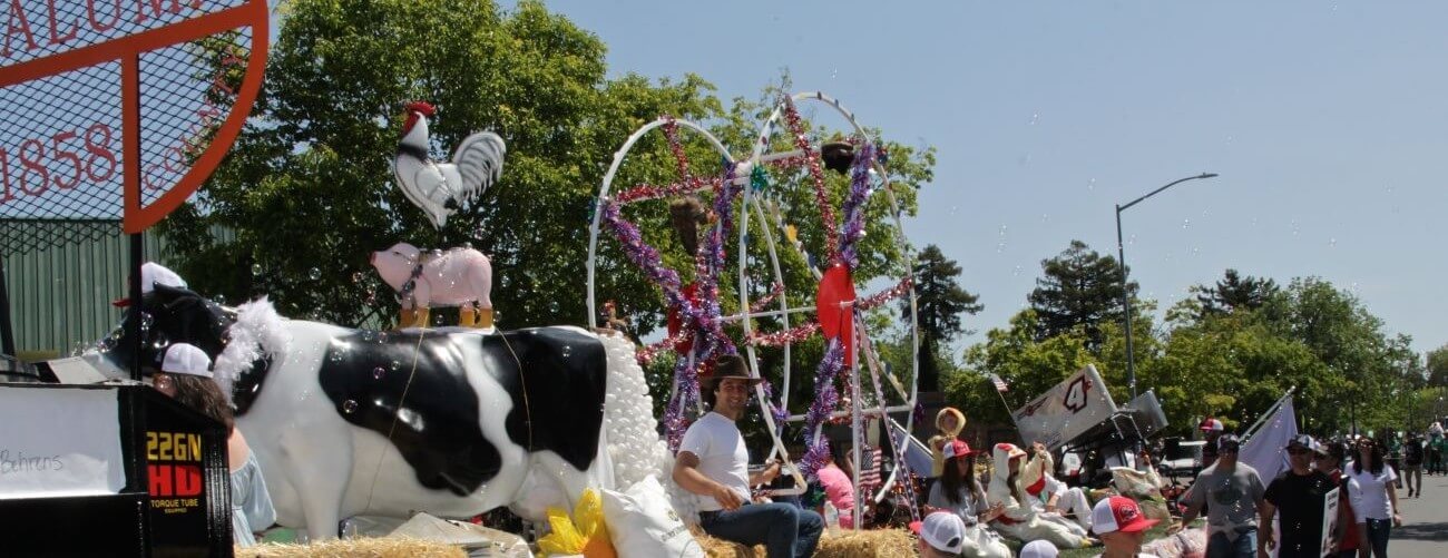 Parade float with cow, pig, and rooster under a petaluma 1858 sign.
