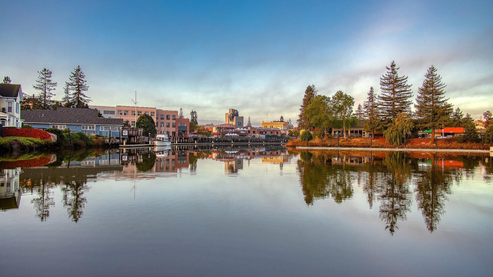Daytime view of downtown Petaluma seen from the river with reflections of building and trees on water.