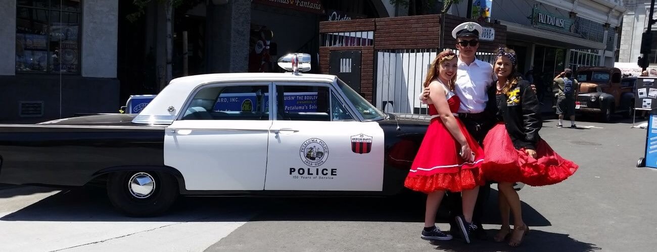 Vintage police cruiser, coast guardsman wearing shades and his arm around 2 poodle-skirted women