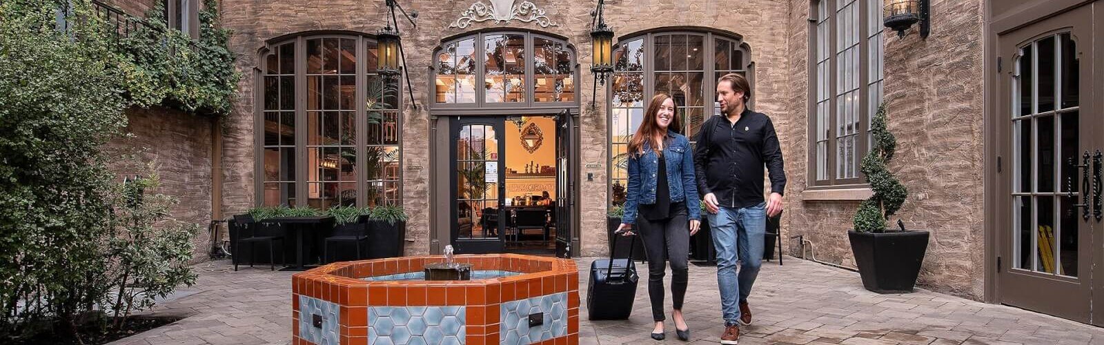 Smiling woman rolling carryon past fountain as she leaves historic hotel with man