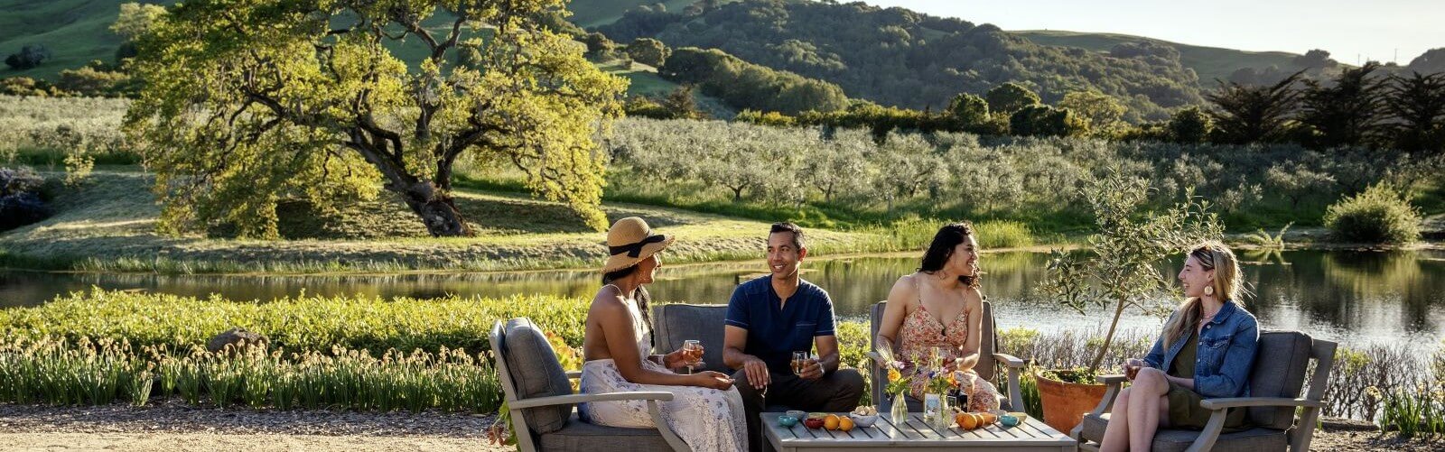 Group of four tasting wine on a summer day with a pond and an oak tree in the background.