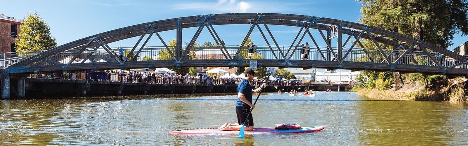 Man on paddleboard on river. Canoes, footbridge, waterfront festival in background