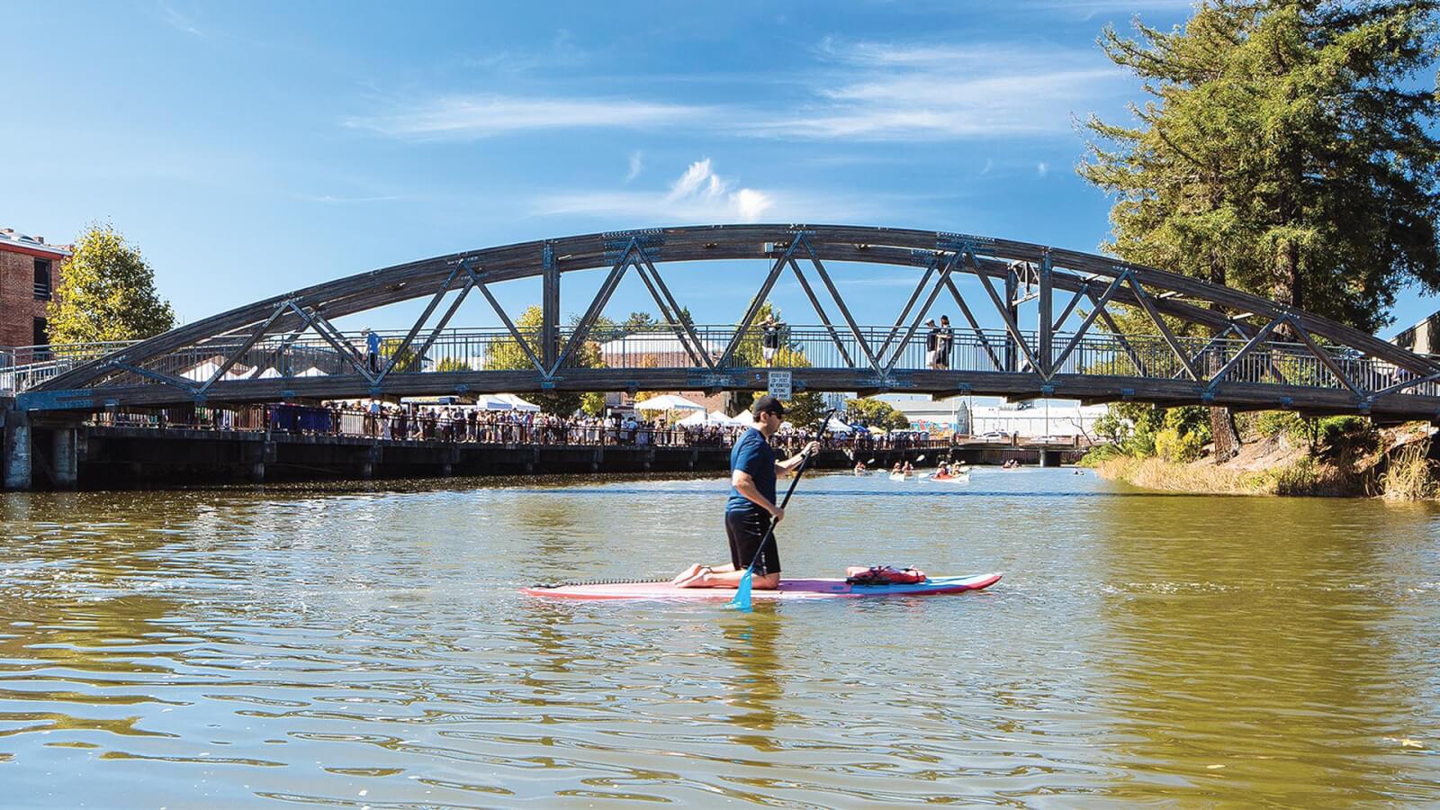 Man on paddleboard on river with footbridge and street fair in background.