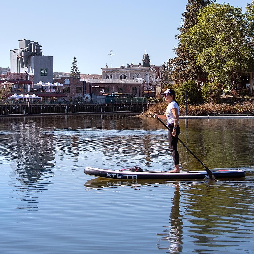 woman on stand up paddleboard on Petaluma river with historic buildings in background.