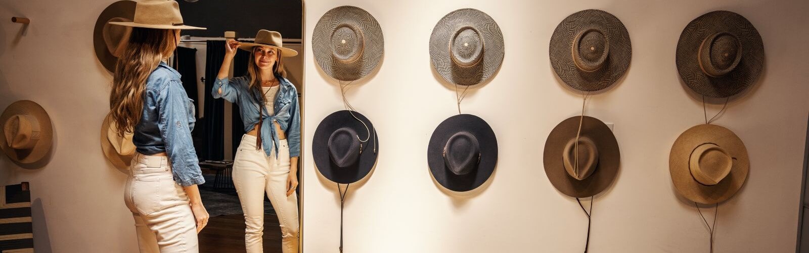Shopping 1 Long-haired woman trying on hat, looking in mirror. Hats on wall.