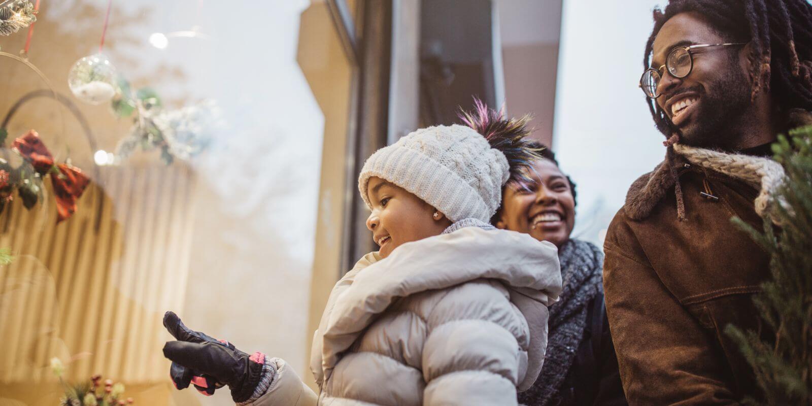 African american family of three holiday shopping. Dad has tree, daughter pointing at display window.